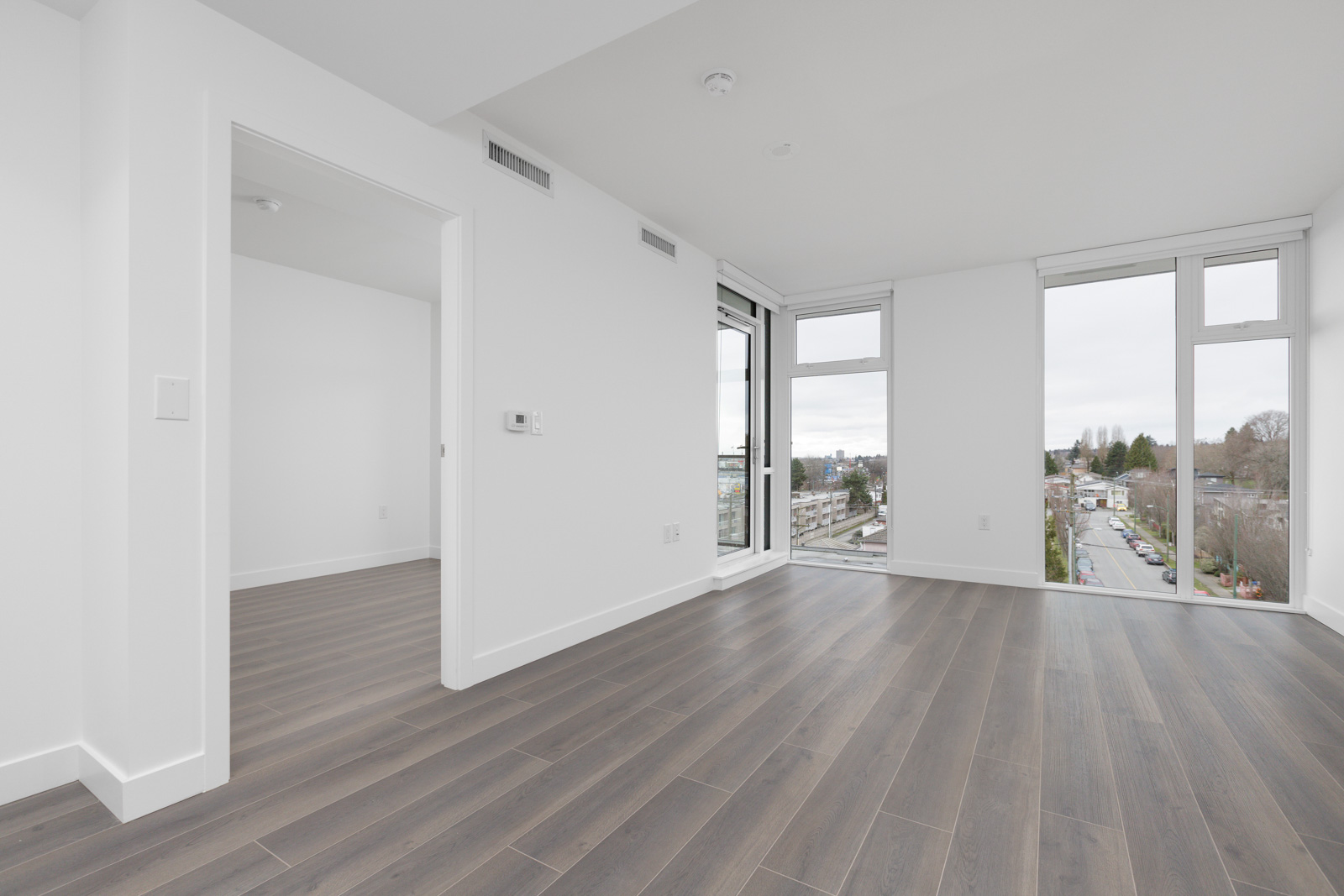 Unfurnished modern apartment room with large windows, white walls, wood flooring, and a doorway leading to another room. Cityscape and greenery visible outside.