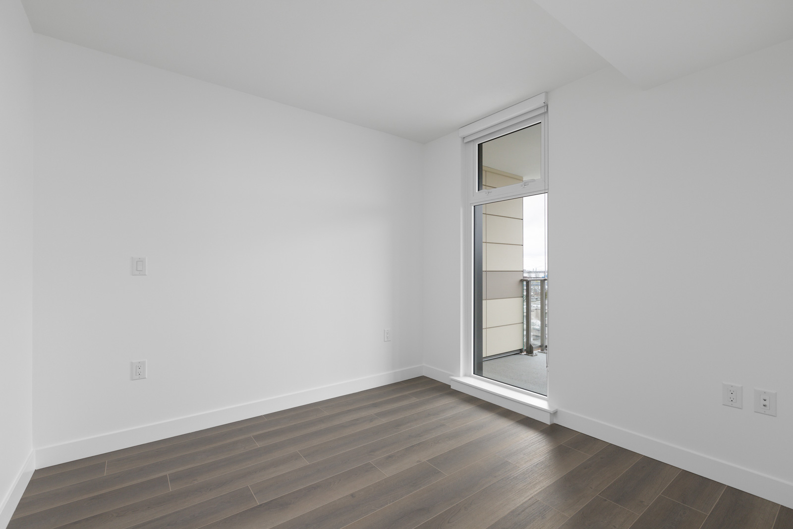 Empty room with white walls, dark wood flooring, and a glass door leading to a small balcony with a partial view outside.