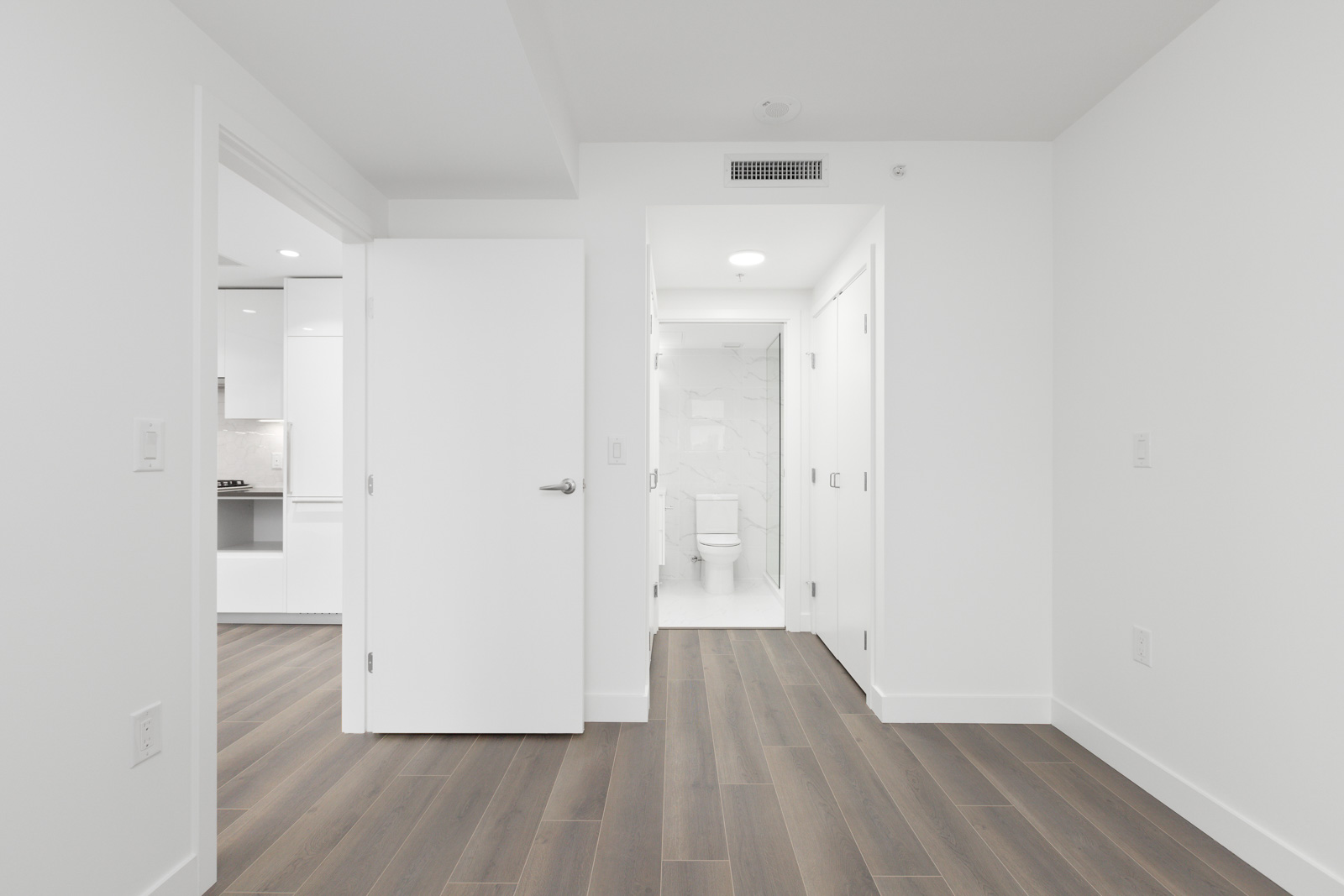 Bright, empty room with wood flooring, white walls, and open doors leading to a kitchen on the left and a bathroom with a visible toilet straight ahead.