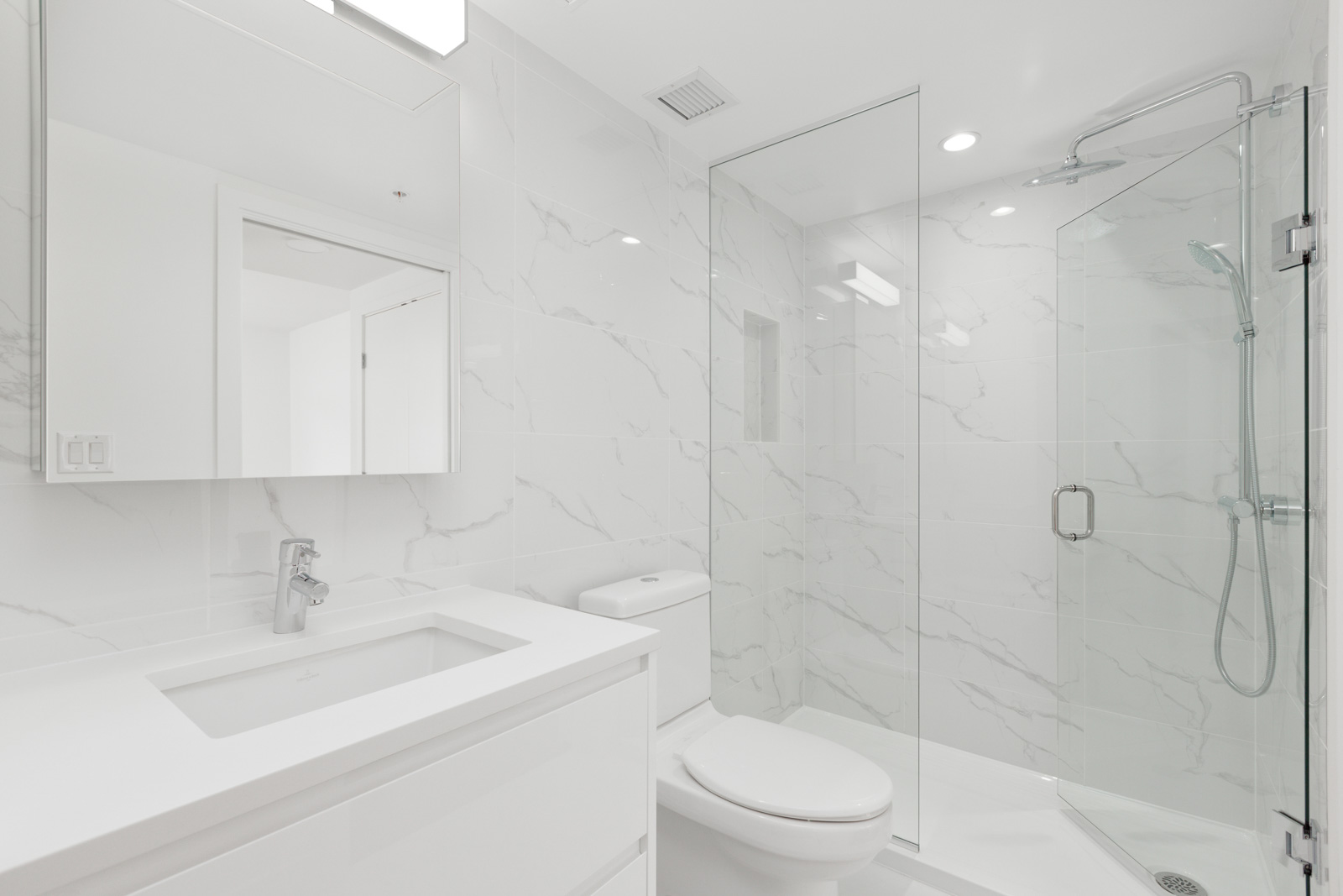 Modern bathroom with white marble tiles, a glass-enclosed shower, a toilet, a sink with an under-mounted basin, and a mirrored cabinet above the vanity.