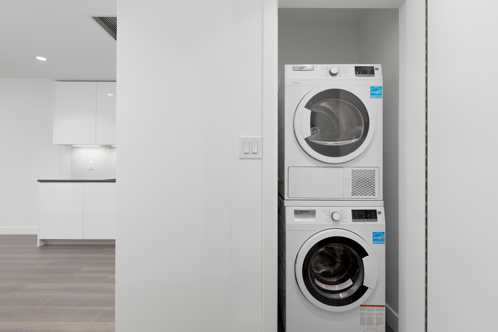 A stacked white washer and dryer set is installed in a small laundry nook next to a modern kitchen with white cabinets and wood flooring.