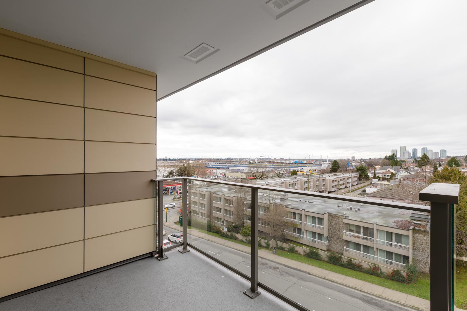 A covered balcony with glass railing overlooks a street, residential buildings, and a city skyline under a cloudy sky.