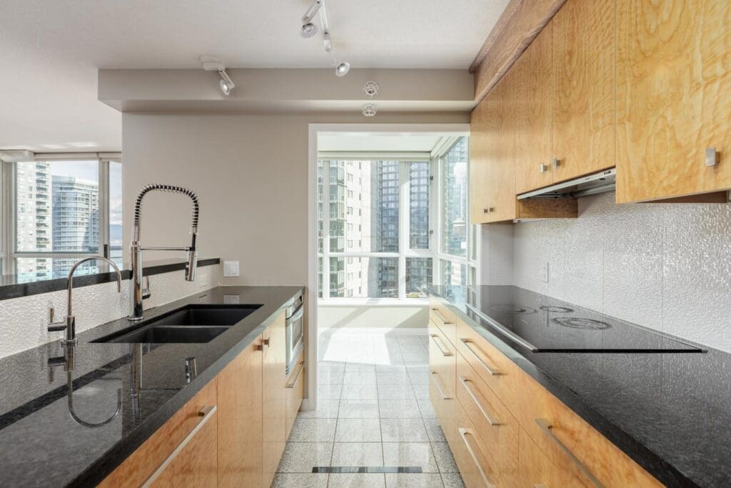 Modern kitchen with light wood cabinets, black countertops, a double sink, electric stove, and large windows overlooking city buildings.
