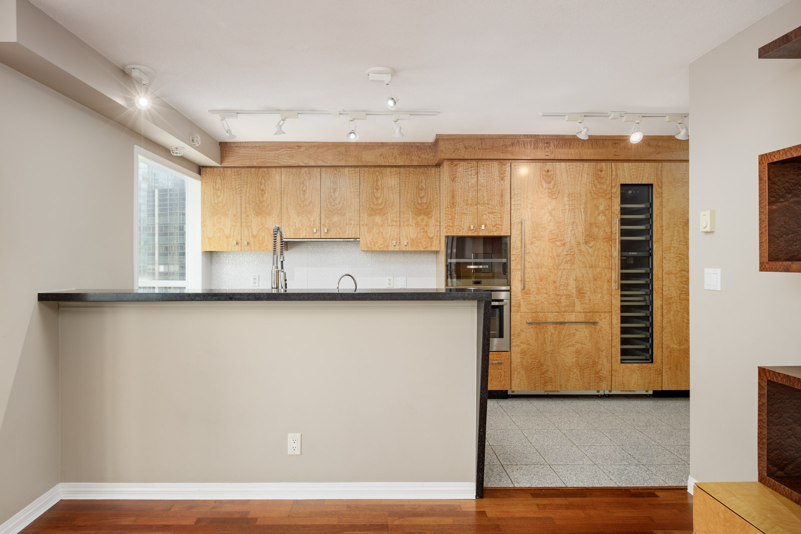 Modern kitchen with wood cabinets, built-in appliances, a wine fridge, a raised breakfast bar, and track lighting. The floor transitions from wood to tile near the kitchen area.
