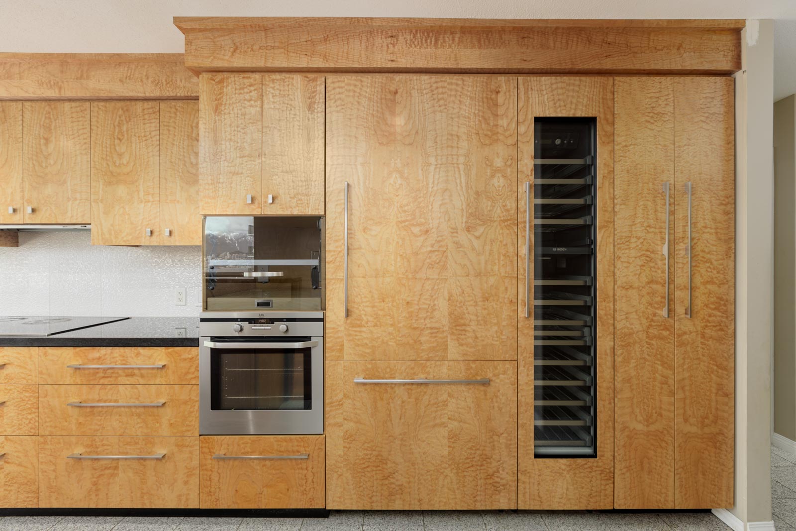 Modern kitchen with light wood cabinets, built-in stainless steel oven, microwave, and a tall glass-front wine fridge, all set against a neutral backsplash.