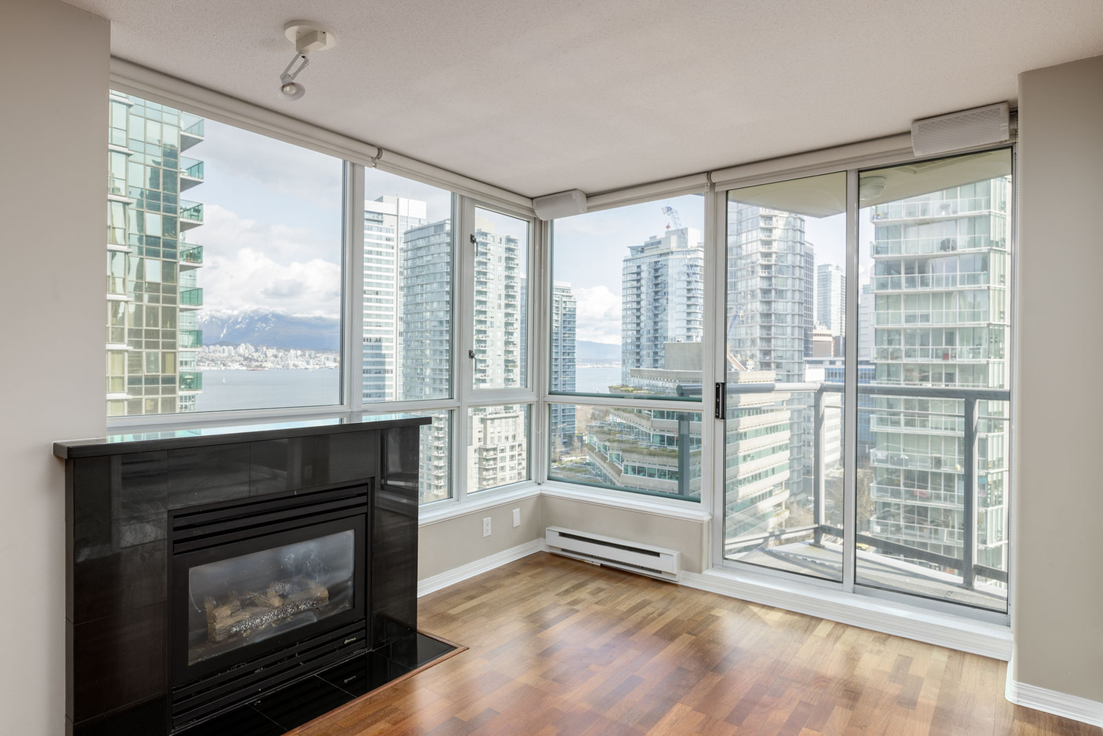 Corner apartment with floor-to-ceiling windows, a black fireplace, hardwood floors, and views of nearby high-rise buildings and distant mountains.