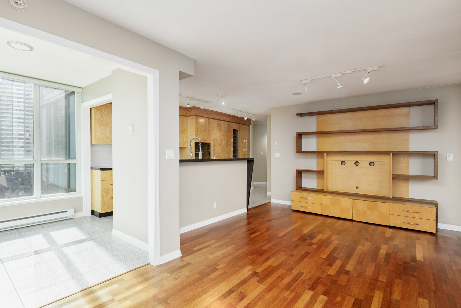 Modern apartment interior with hardwood floors, built-in wooden shelves, a kitchen area with light wood cabinetry, and large windows providing natural light.