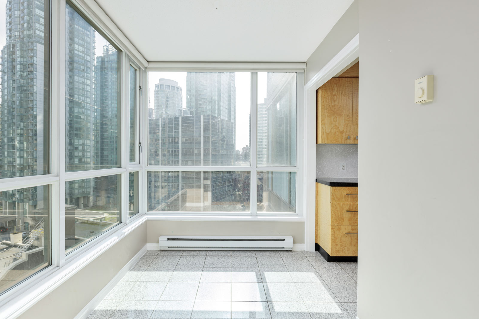 Bright corner room with large floor-to-ceiling windows overlooking city buildings, tiled floor, and partial view of a kitchen with wooden cabinets.