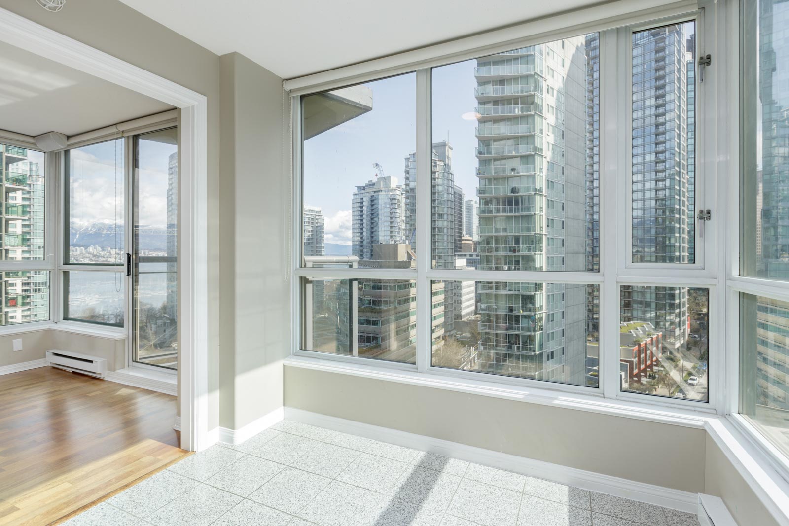 A bright, empty corner room with large windows overlooks modern high-rise buildings and a partial view of water in an urban setting.