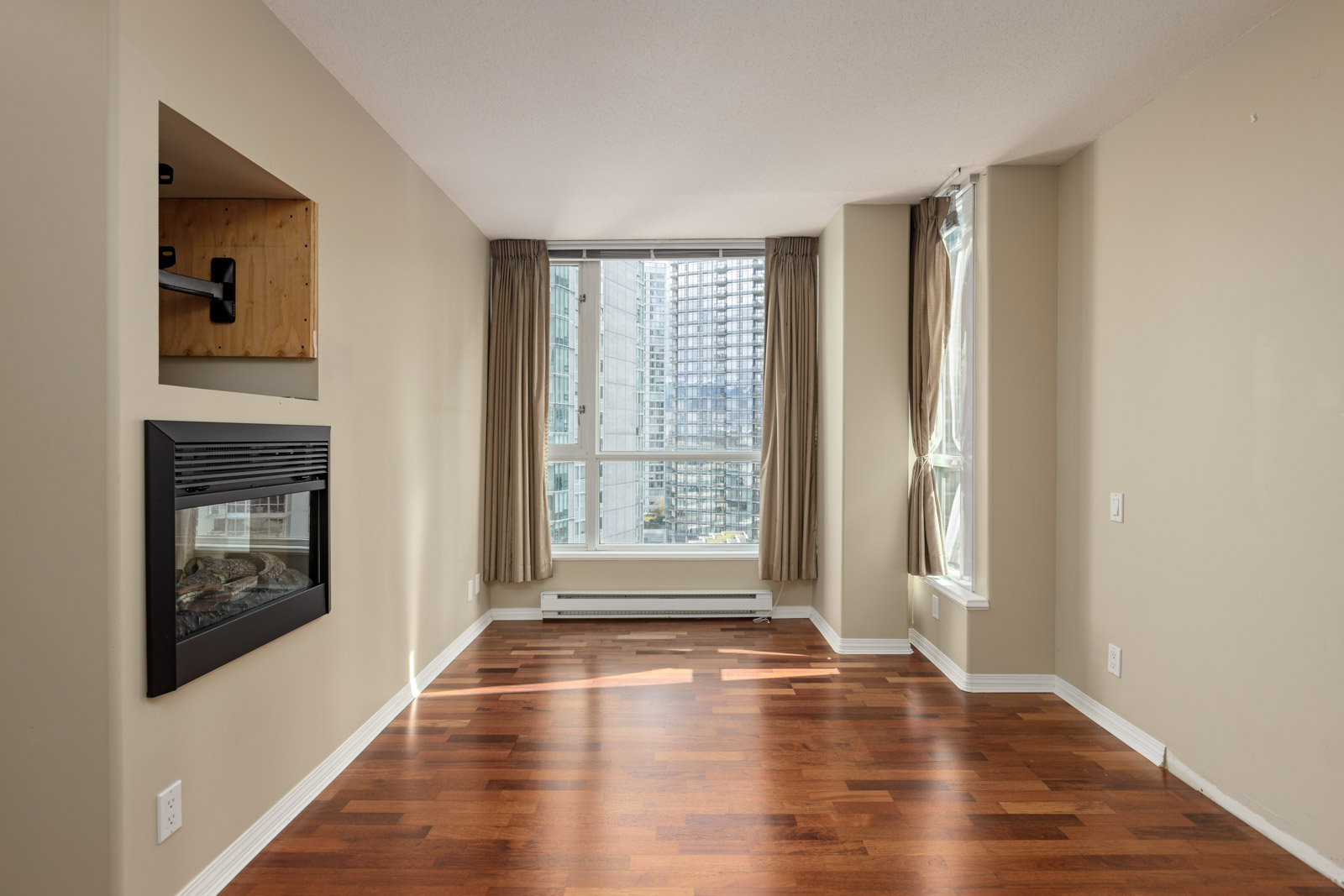 Unfurnished living room with hardwood floor, built-in fireplace, large windows with beige curtains, and a cityscape view.