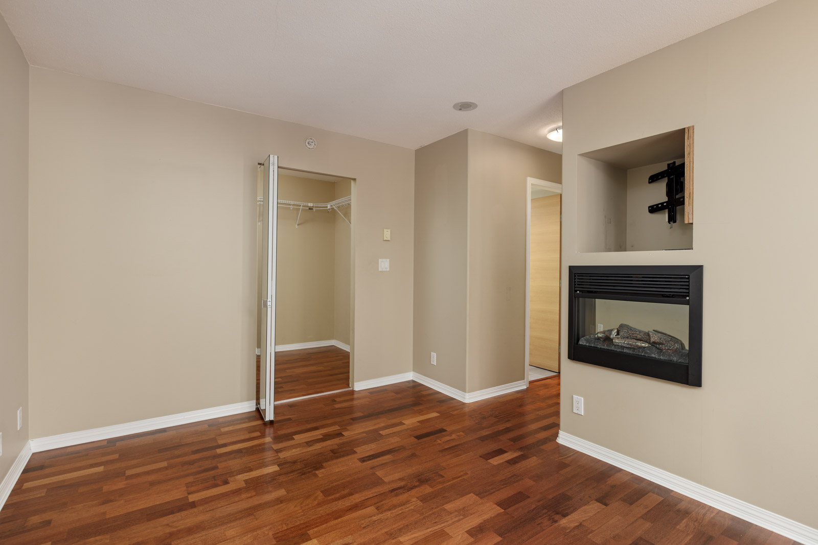Empty room with light beige walls, a closet with mirrored doors, wood flooring, a wall-mounted electric fireplace, and an open doorway leading to a hallway.