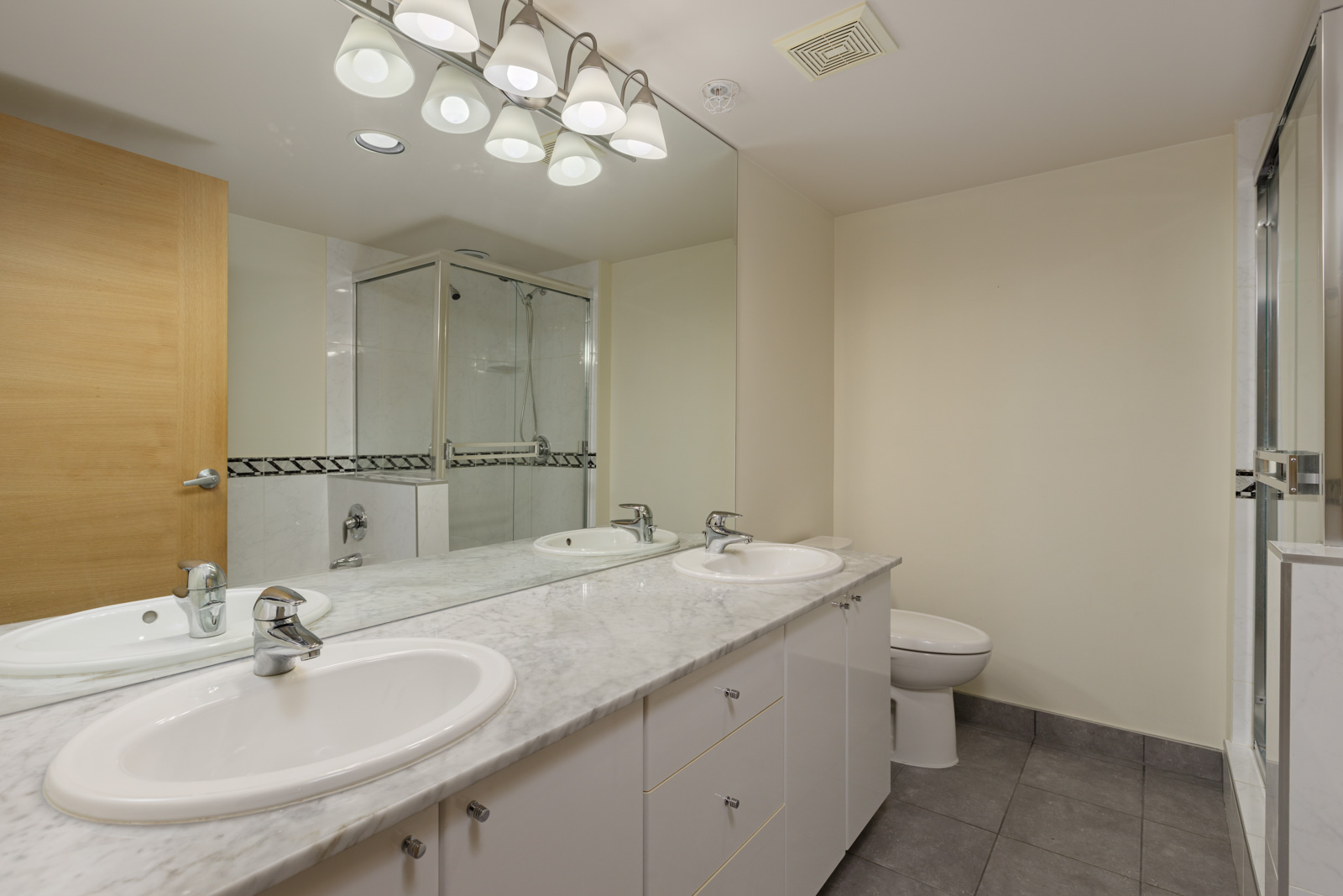 Modern bathroom with a large mirror, double sinks, marble countertop, toilet, glass-enclosed shower, tiled floor, and bright overhead lighting.