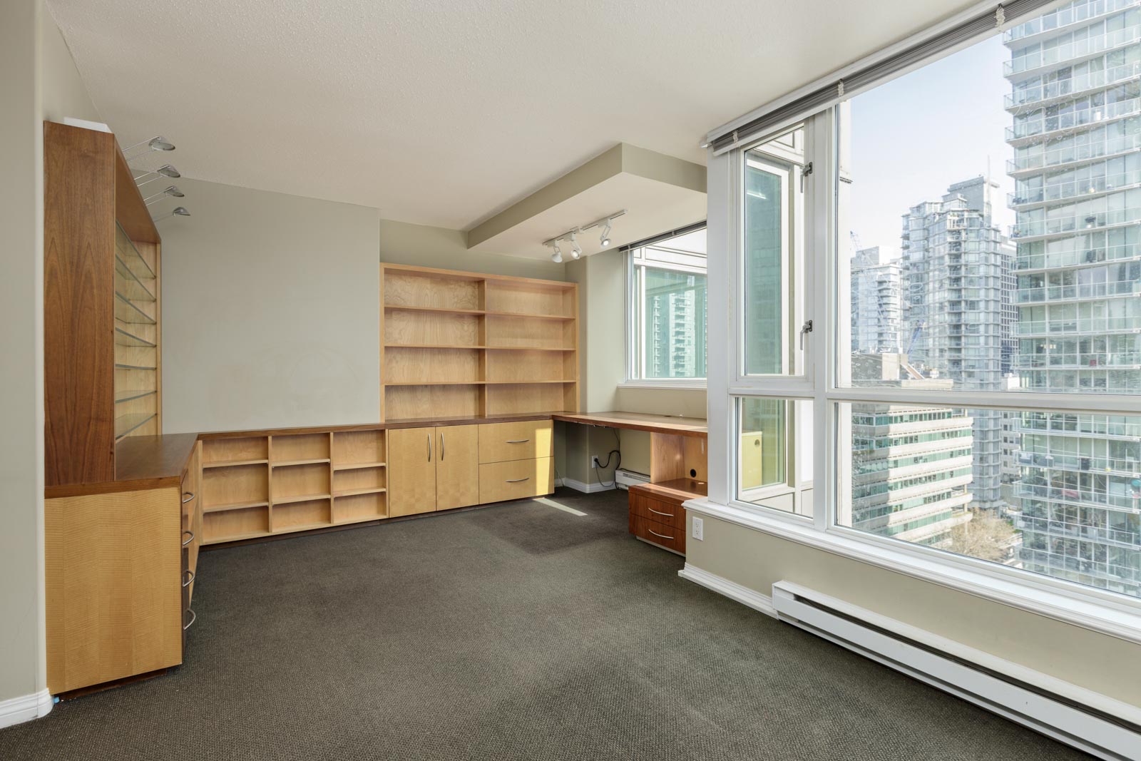 Bright empty office space with built-in wooden shelves and desk, large windows, carpet flooring, and city high-rise buildings visible outside.