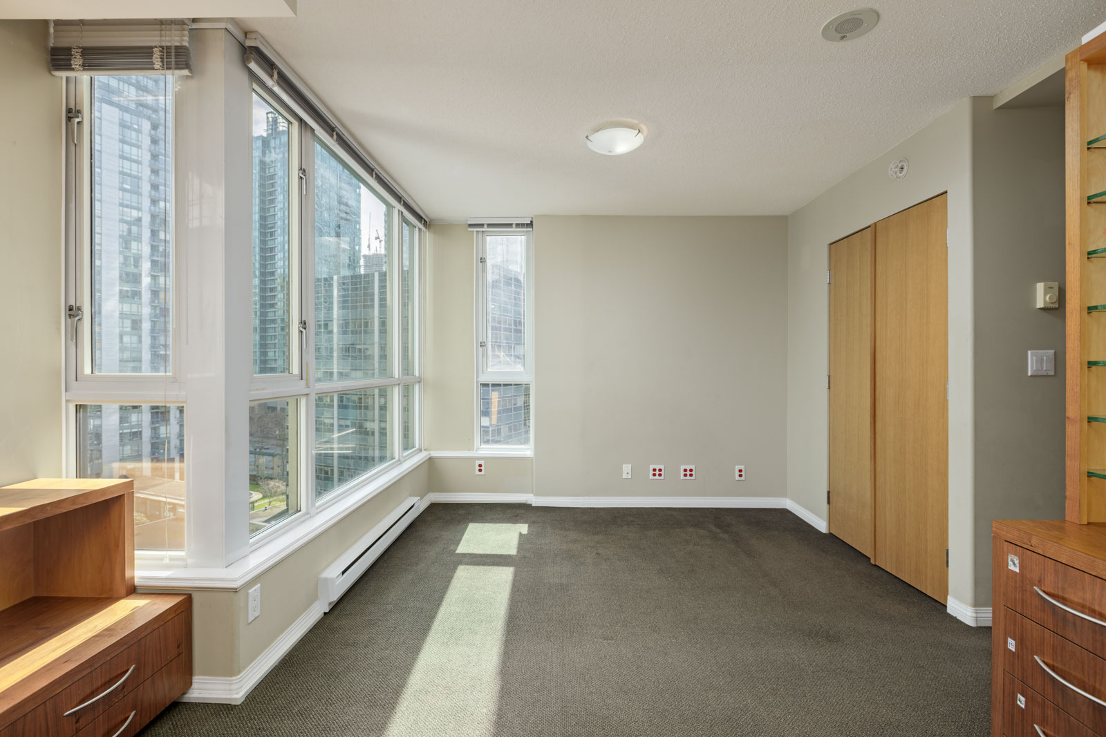 Unfurnished corner room with large floor-to-ceiling windows, beige walls, grey carpet, wood furniture, and city buildings visible outside.