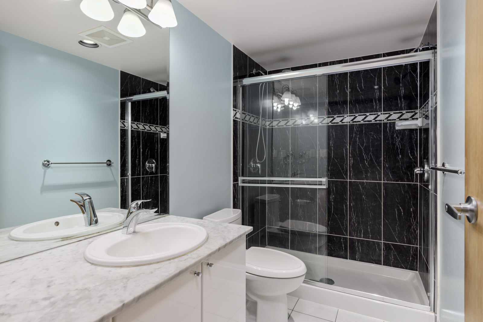 Modern bathroom with a marble countertop, white sink, toilet, and a glass-enclosed shower with black tile walls and decorative border.