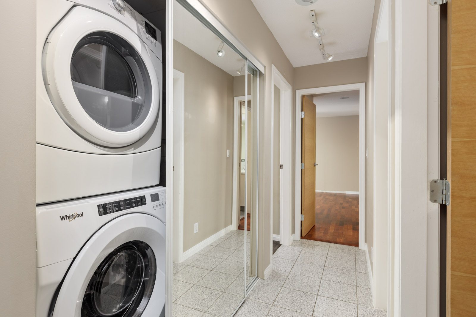 Stacked Whirlpool washer and dryer are next to mirrored closet doors in a hallway with tile flooring and multiple doorways.