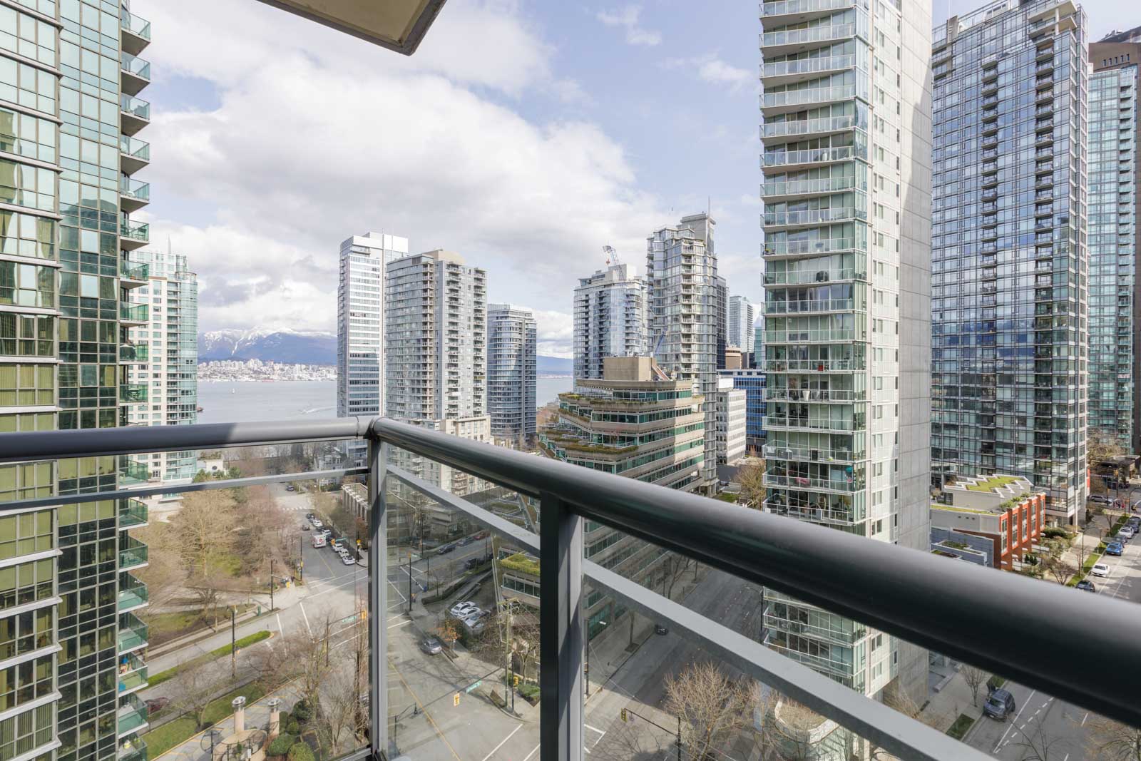View from a balcony overlooking a cityscape with tall modern buildings, tree-lined streets, and distant water and mountains under a partly cloudy sky.