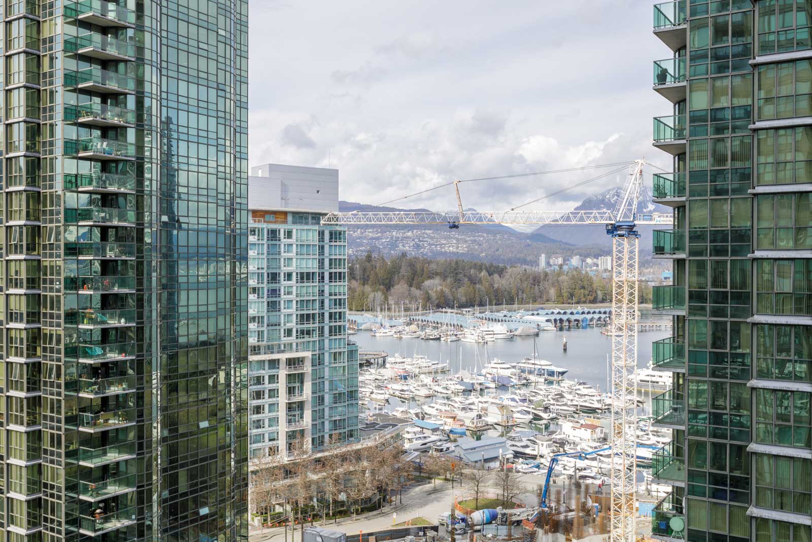 High-rise glass buildings frame a marina filled with boats; a construction crane and mountains are visible in the background under a cloudy sky.