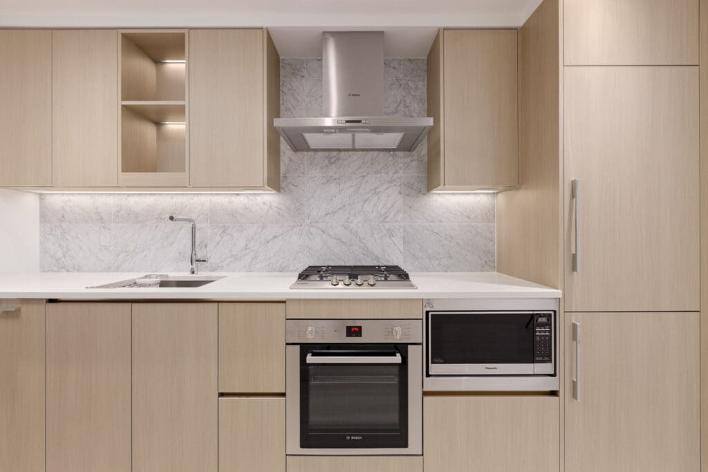 Modern kitchen with light wood cabinets, white countertops, built-in oven, microwave, gas cooktop, range hood, and marble backsplash.