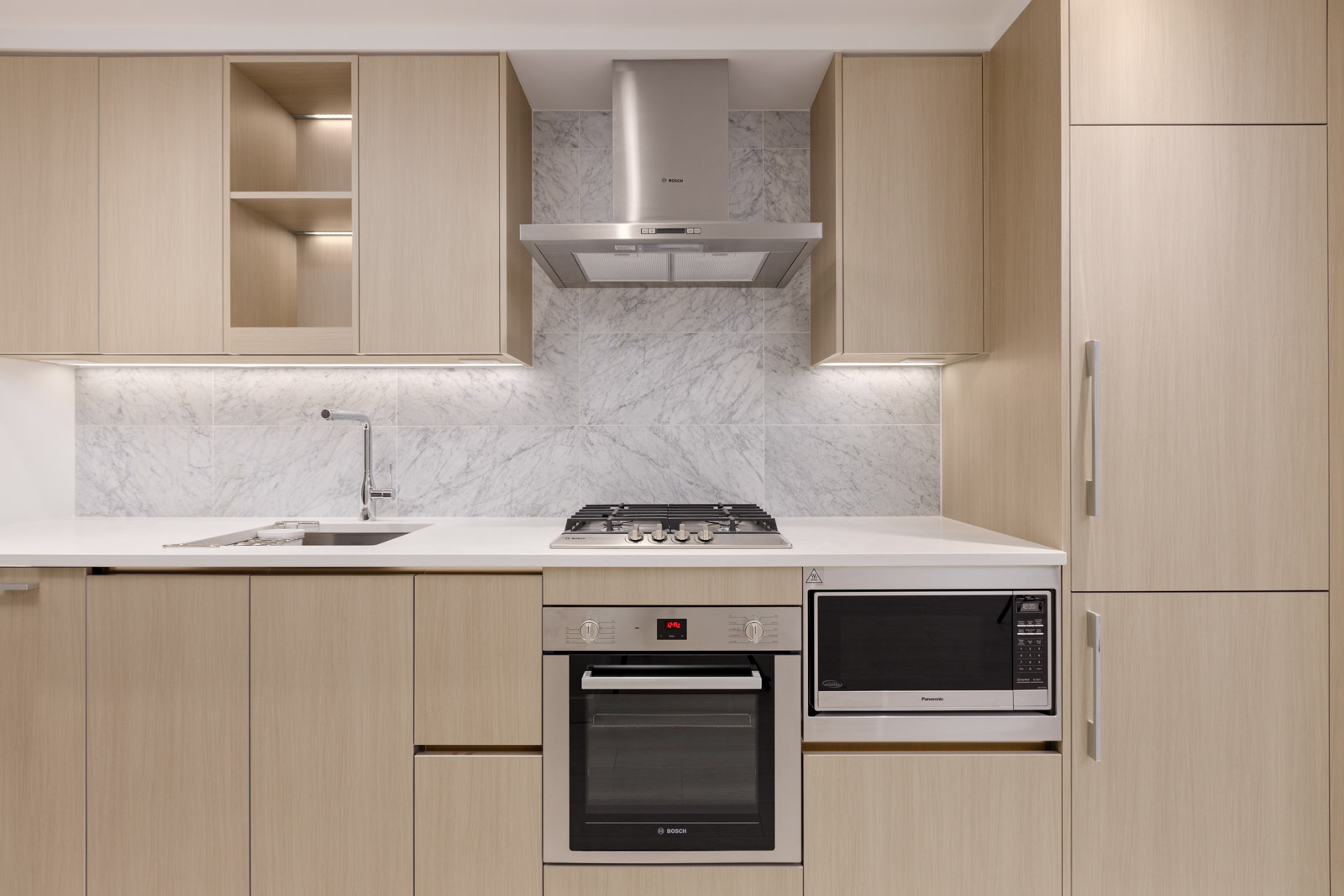 Modern kitchen with light wood cabinets, white countertops, built-in oven, microwave, gas cooktop, range hood, and marble backsplash.