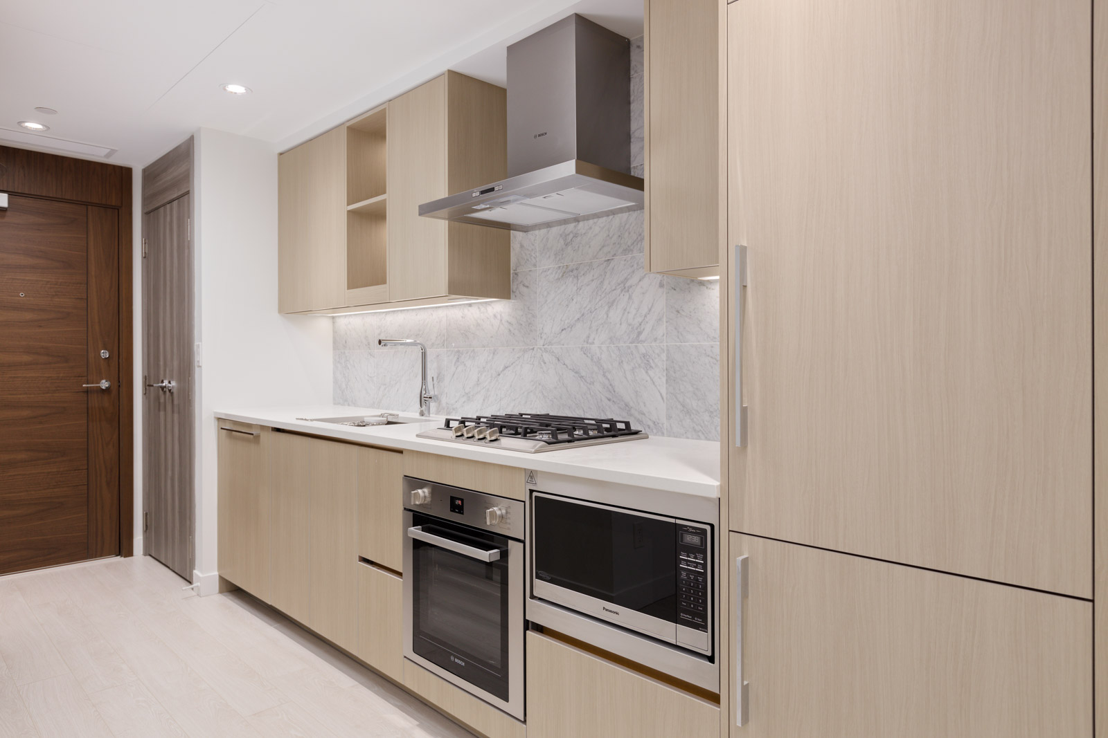 Modern kitchen with light wood cabinets, built-in oven and microwave, gas cooktop, stainless steel range hood, and marble backsplash.