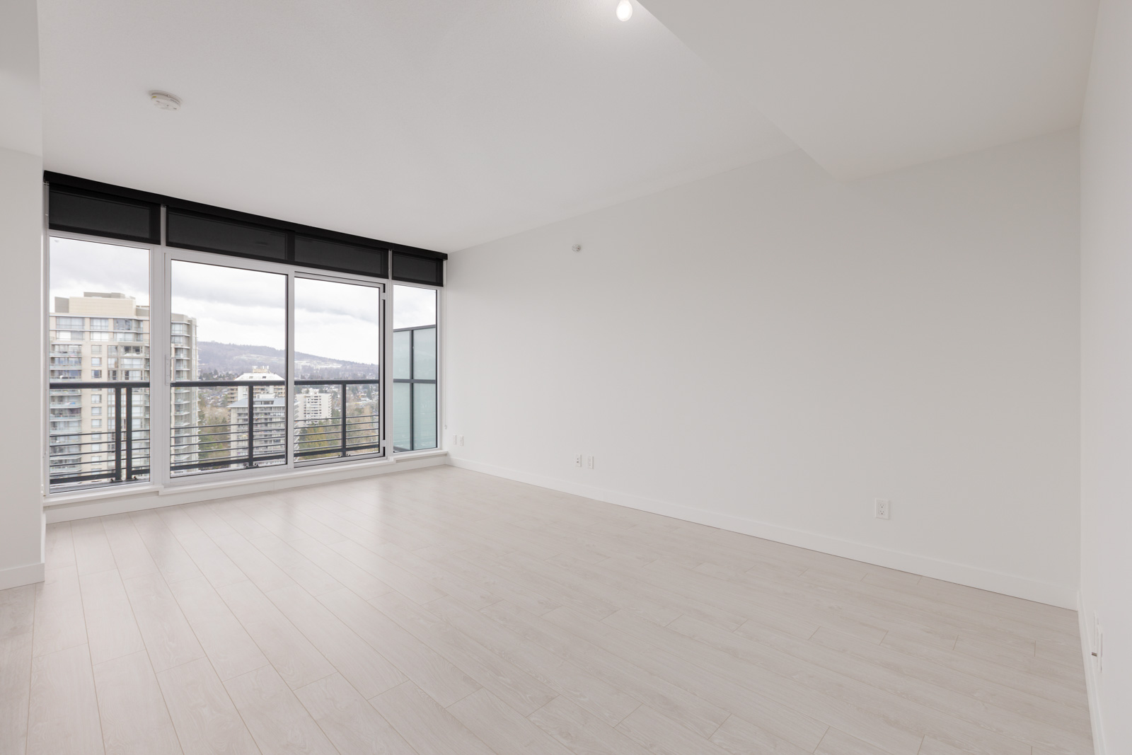 Unfurnished, empty room with light wood floors, white walls, and large floor-to-ceiling windows overlooking a cityscape with mountains in the background.