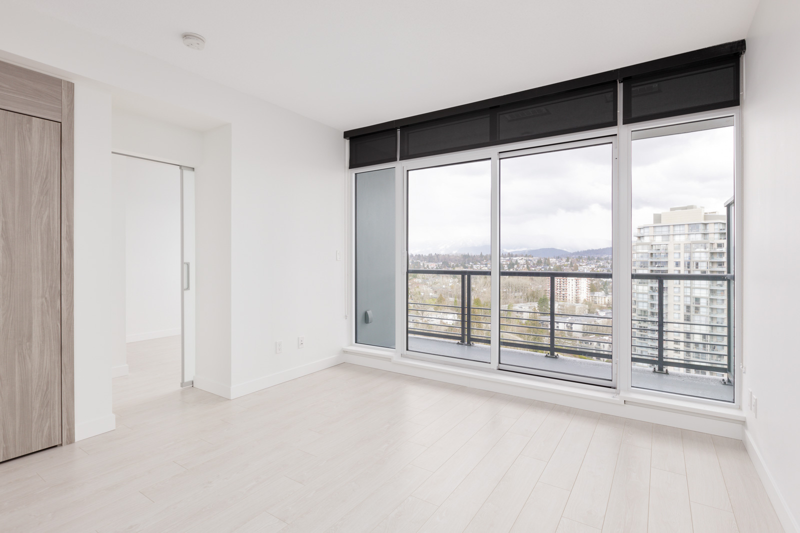Empty modern apartment room with light wood flooring, large windows, sliding glass door, and a view of city buildings and trees outside.
