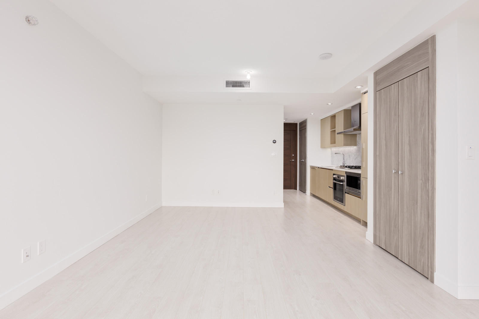 Unfurnished modern apartment with light-colored flooring, a kitchen with wood cabinets on the right, and a closed door at the far end. Walls and ceiling are white.