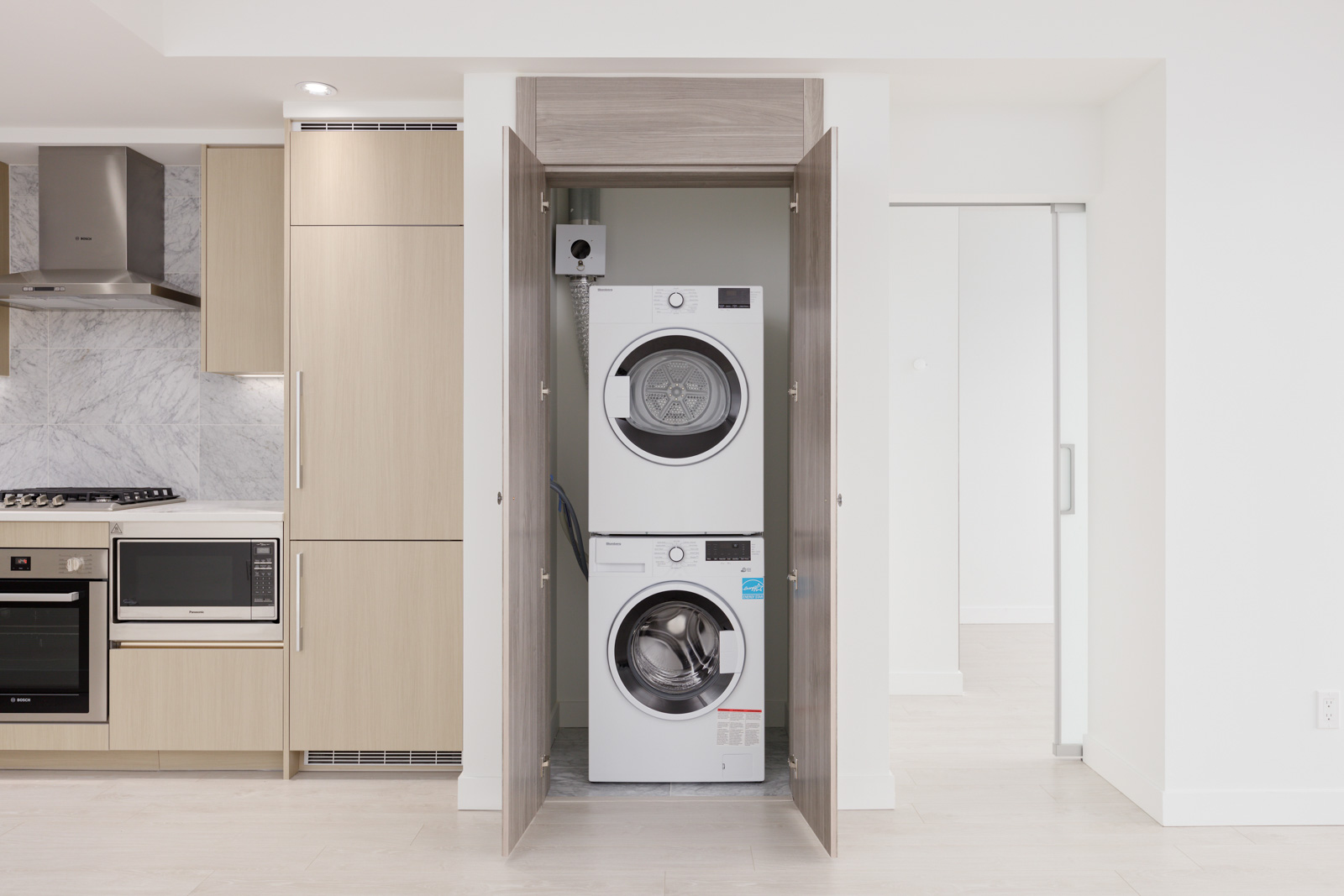 A stacked washer and dryer are enclosed in a wooden closet next to a modern kitchen with light wood cabinets and stainless steel appliances.
