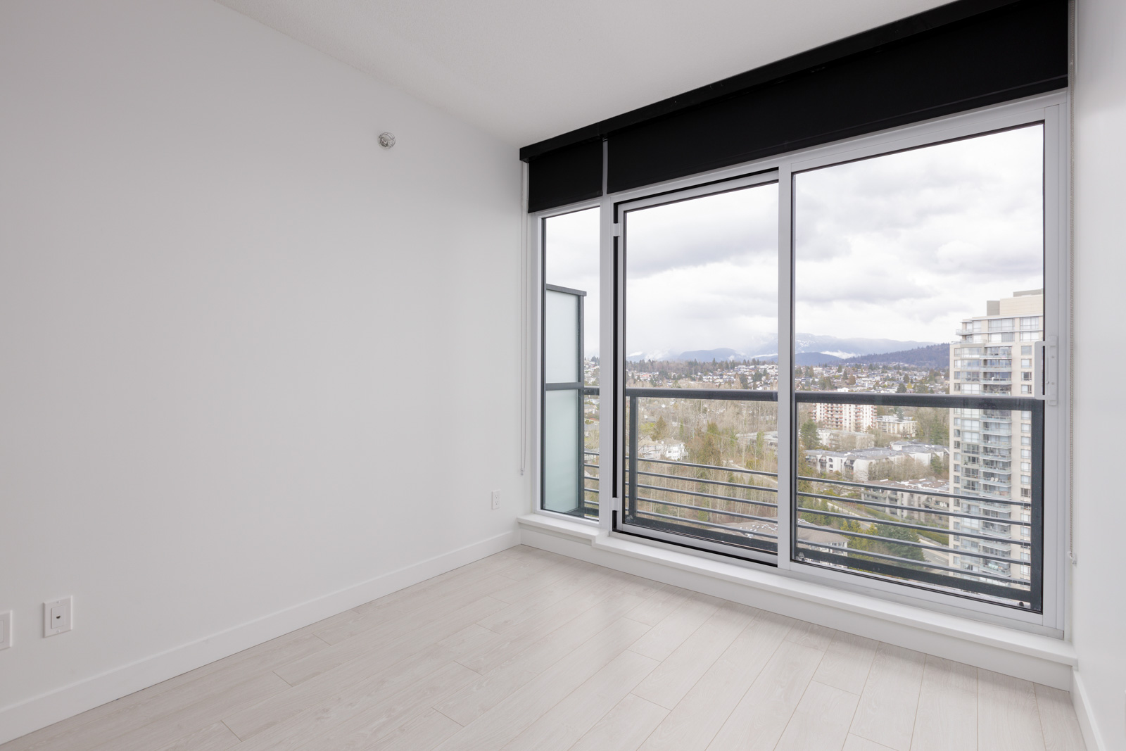 Empty modern room with light wood flooring, white walls, and large floor-to-ceiling windows offering a cityscape and mountain view under a cloudy sky.