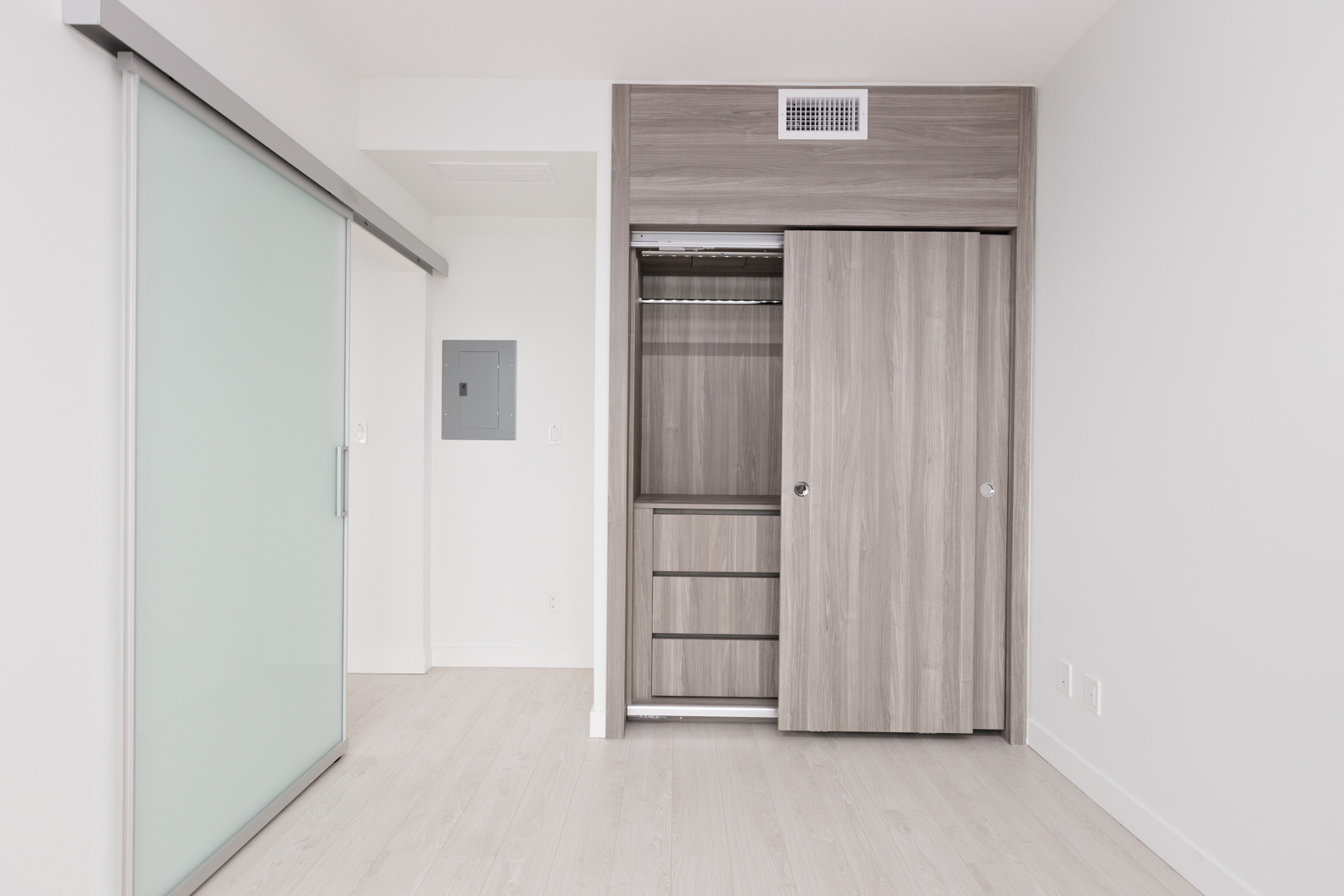 A modern, empty room with light wood flooring, a sliding glass door on the left, and an open built-in closet with drawers and hanging space on the right.