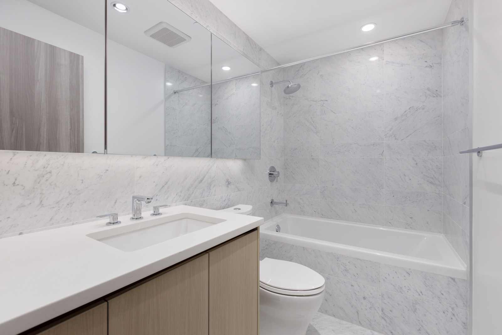 Modern bathroom with marble walls, built-in bathtub with shower, white countertop sink, toilet, large mirror, and recessed lighting.