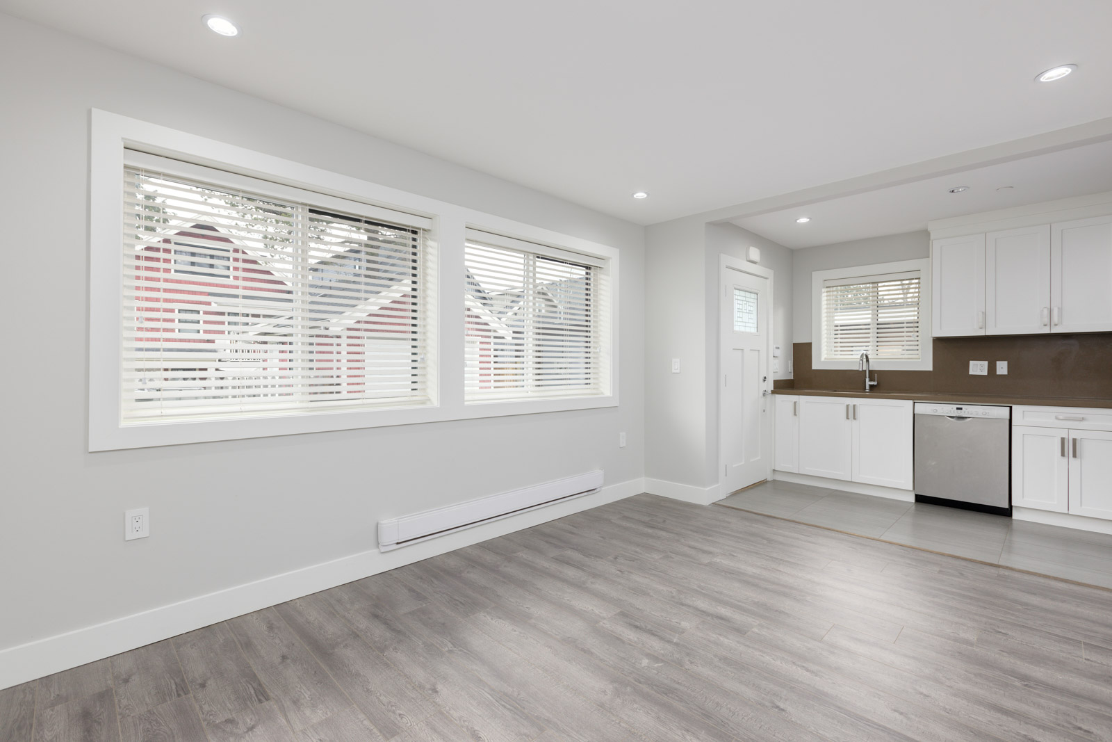 Modern, unfurnished open-concept room with light gray walls, large windows with blinds, wood-style flooring, and a white kitchen area with stainless steel appliances.