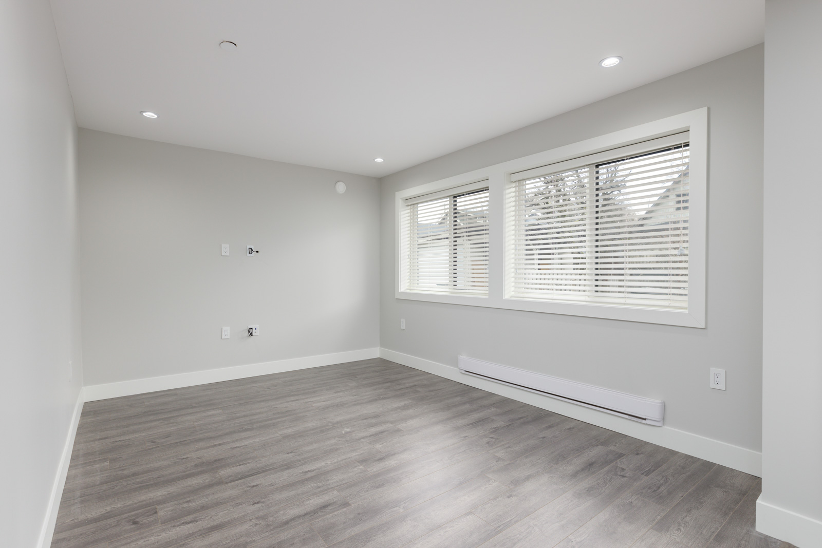 Empty room with light gray walls, large windows with blinds, recessed ceiling lights, and gray wood flooring. Electrical outlets and a baseboard heater are visible.