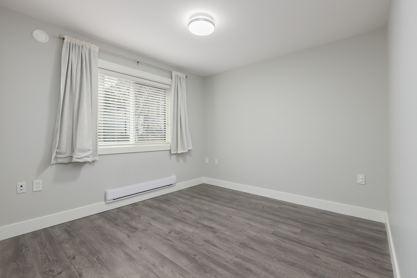 Empty room with light gray walls, wood-style flooring, a window with white blinds and curtains, and a ceiling light.