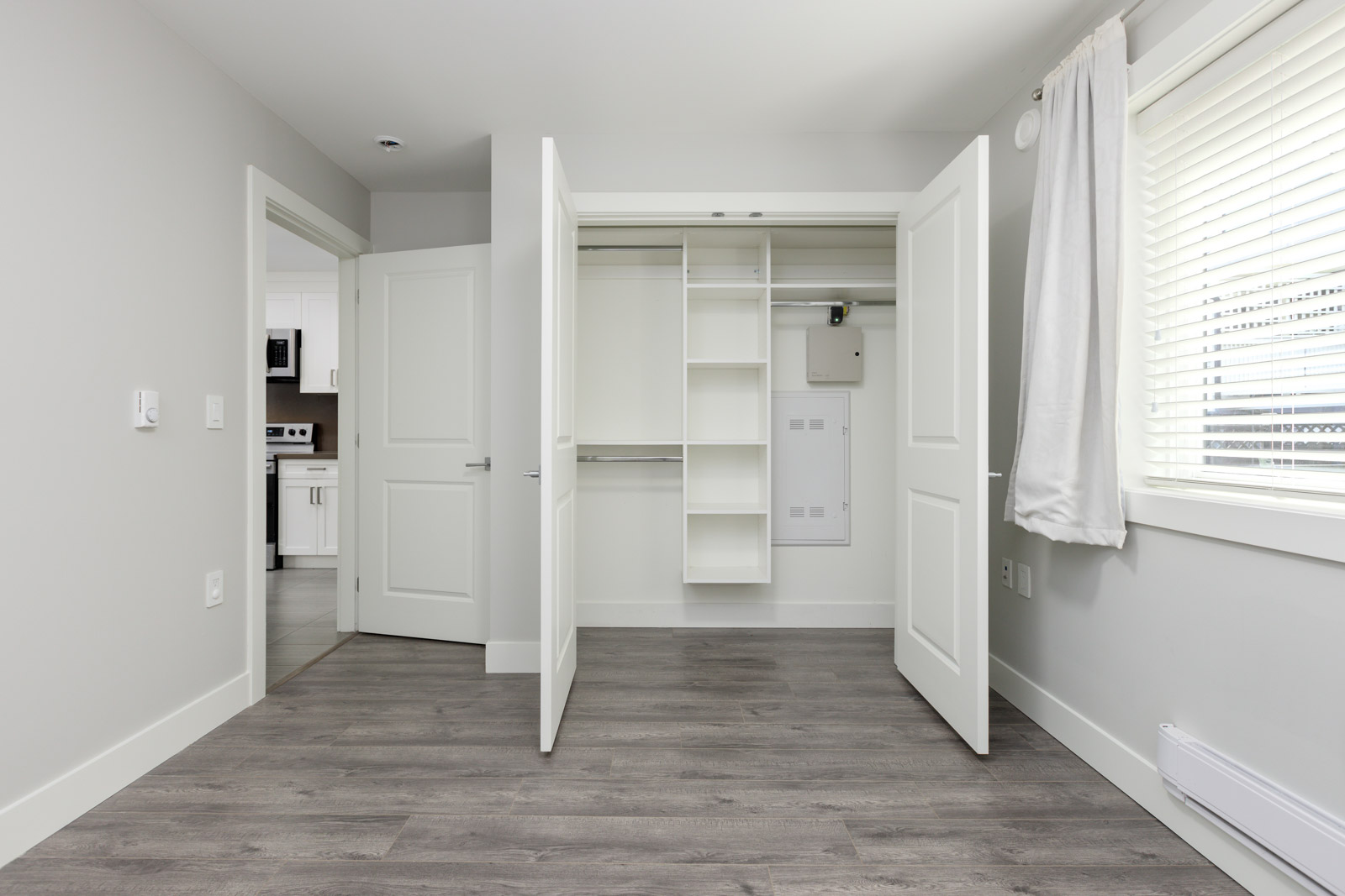 Empty room with open closet doors showing built-in shelves, gray wood flooring, white walls, a window with blinds, and a view into a kitchen area.