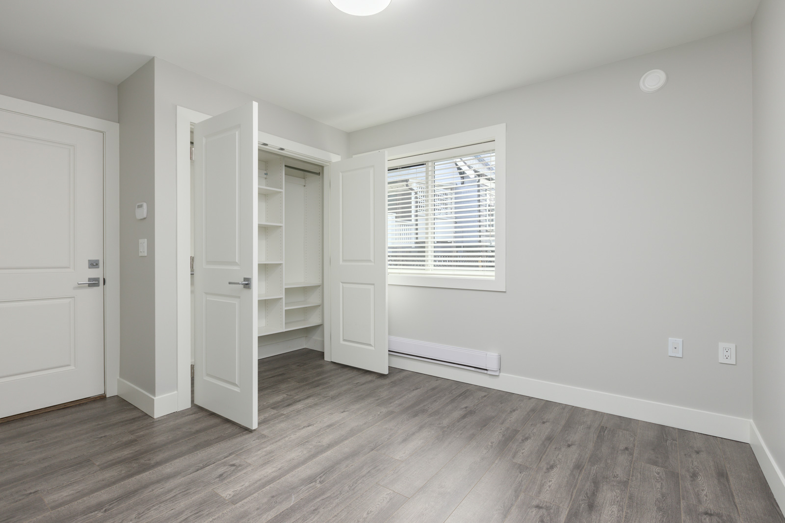 Empty room with light gray walls, wood-patterned flooring, open double closet doors, a window with blinds, and white trim.