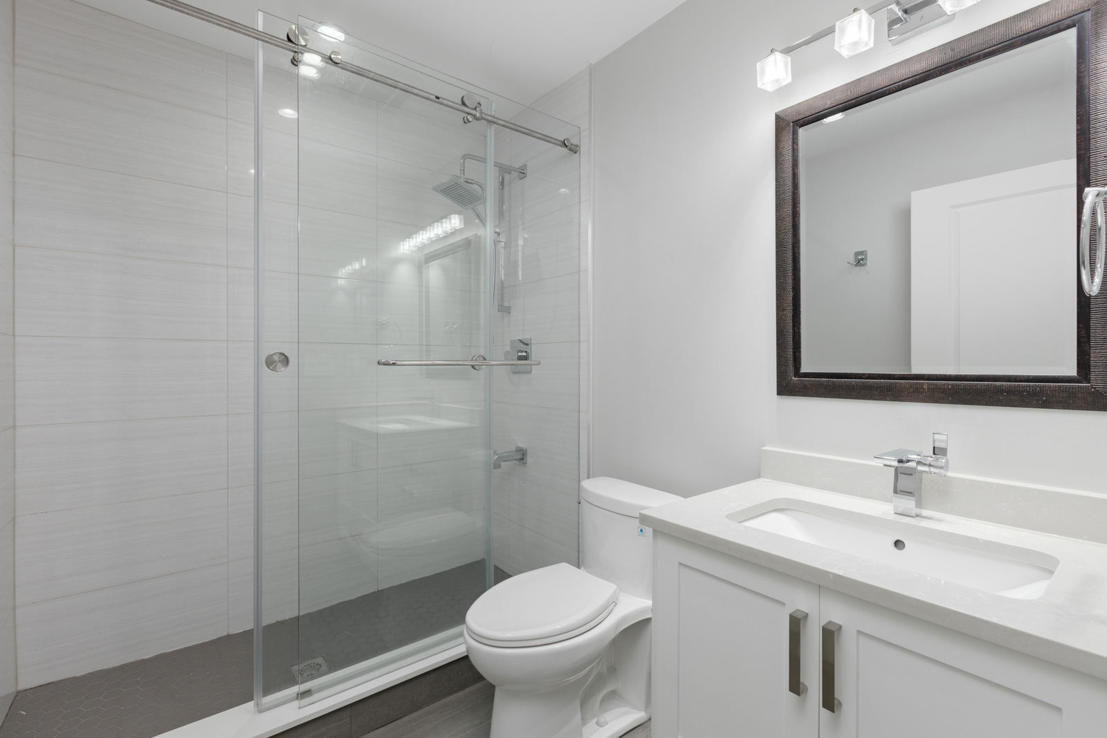Modern bathroom with a glass-enclosed shower, white vanity with sink, large mirror, and a toilet. The walls and fixtures are clean and minimalistic.