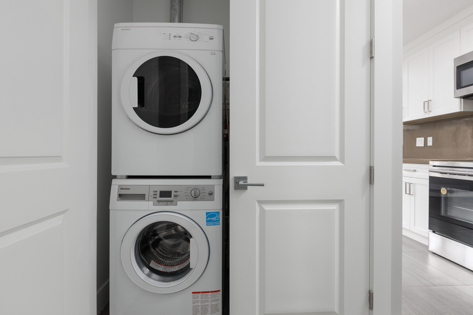A stacked washer and dryer are installed in a small laundry closet with white double doors, next to a modern kitchen.