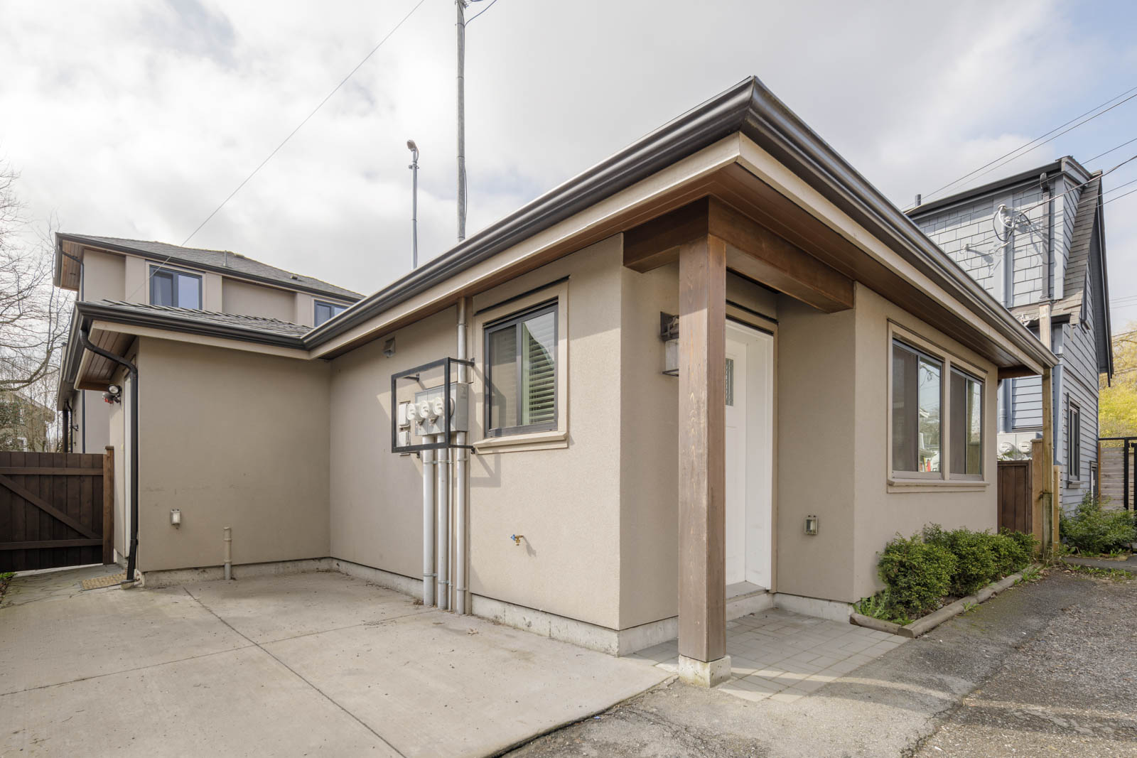 Single-story beige house with flat roof, small covered entrance, concrete driveway, utility meters on the wall, and minimal landscaping.