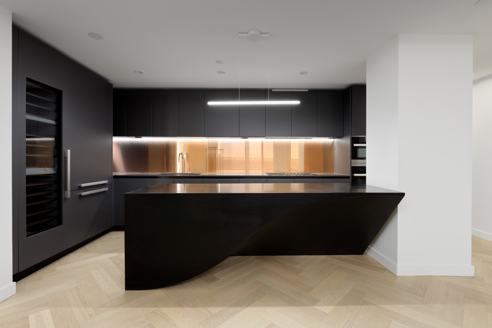 Modern kitchen with matte black cabinets, a black island featuring a curved design, built-in appliances, wood herringbone floor, and warm under-cabinet lighting.