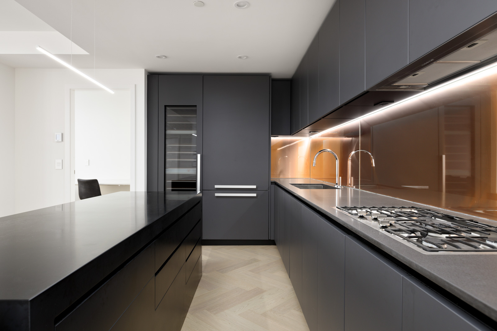 Modern kitchen with dark cabinetry, black countertops, built-in wine fridge, gas cooktop, and a copper-colored backsplash under under-cabinet lighting.