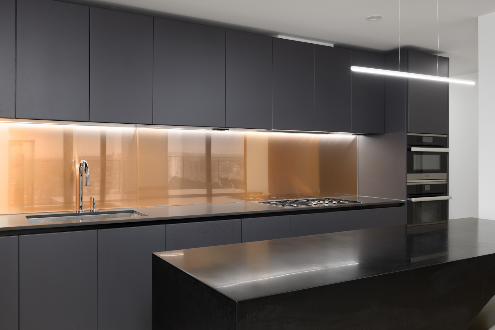 Modern kitchen with dark cabinets, built-in appliances, an island, and a long horizontal light fixture above. The backsplash is a reflective bronze glass.