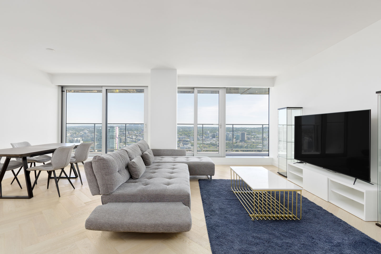 Modern living room with gray sectional sofa, dining table, large TV, gold coffee table, and floor-to-ceiling windows overlooking a cityscape.