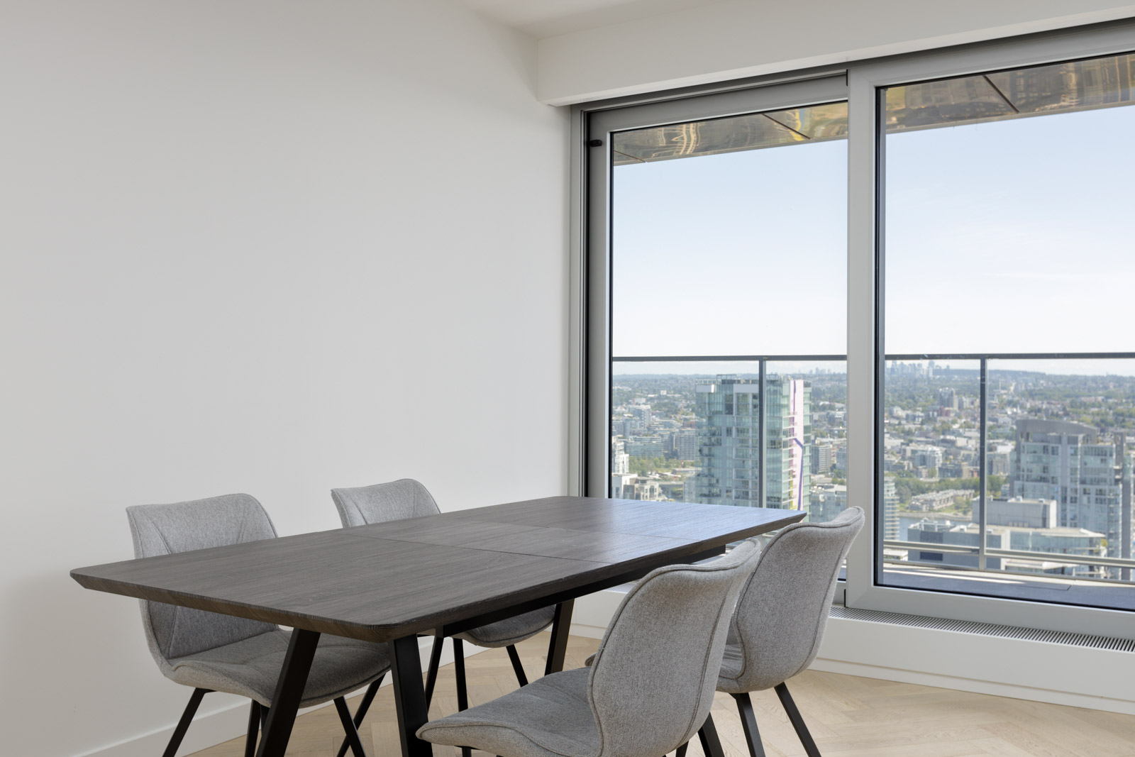 A modern dining table with four gray chairs is positioned by large windows overlooking a cityscape from a high-rise apartment.