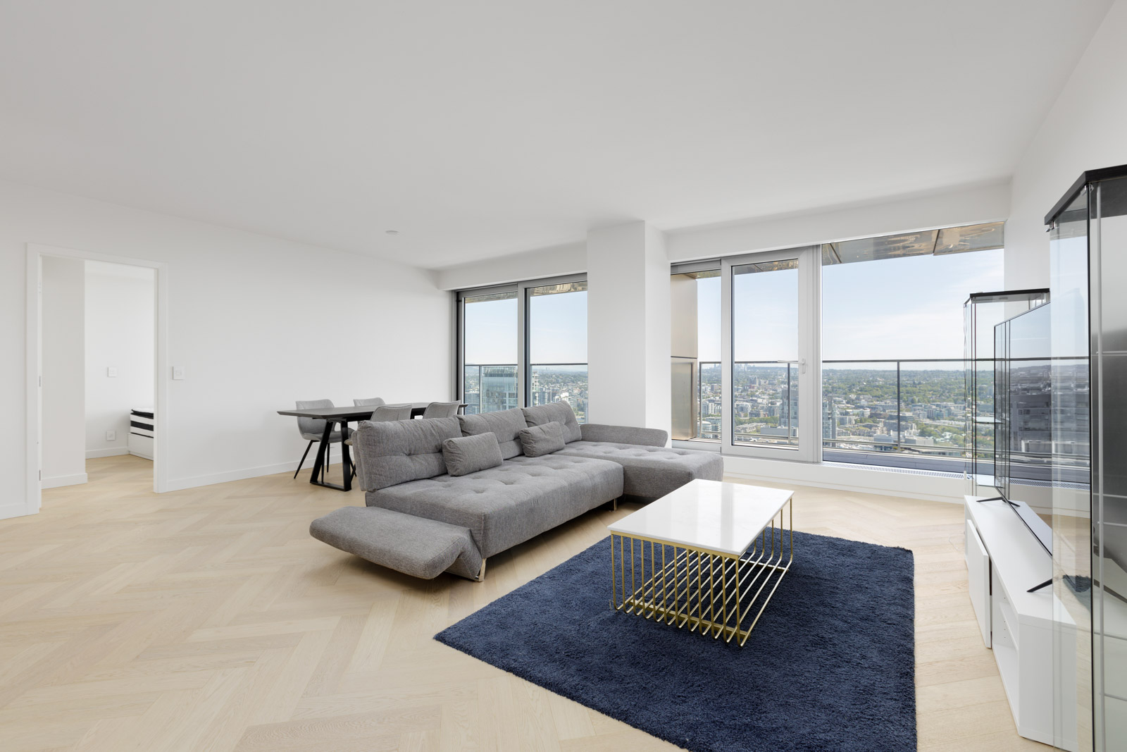 A modern, minimalist living room with a gray sectional sofa, gold coffee table, navy rug, glass display cabinet, dining table, and large windows overlooking a cityscape.
