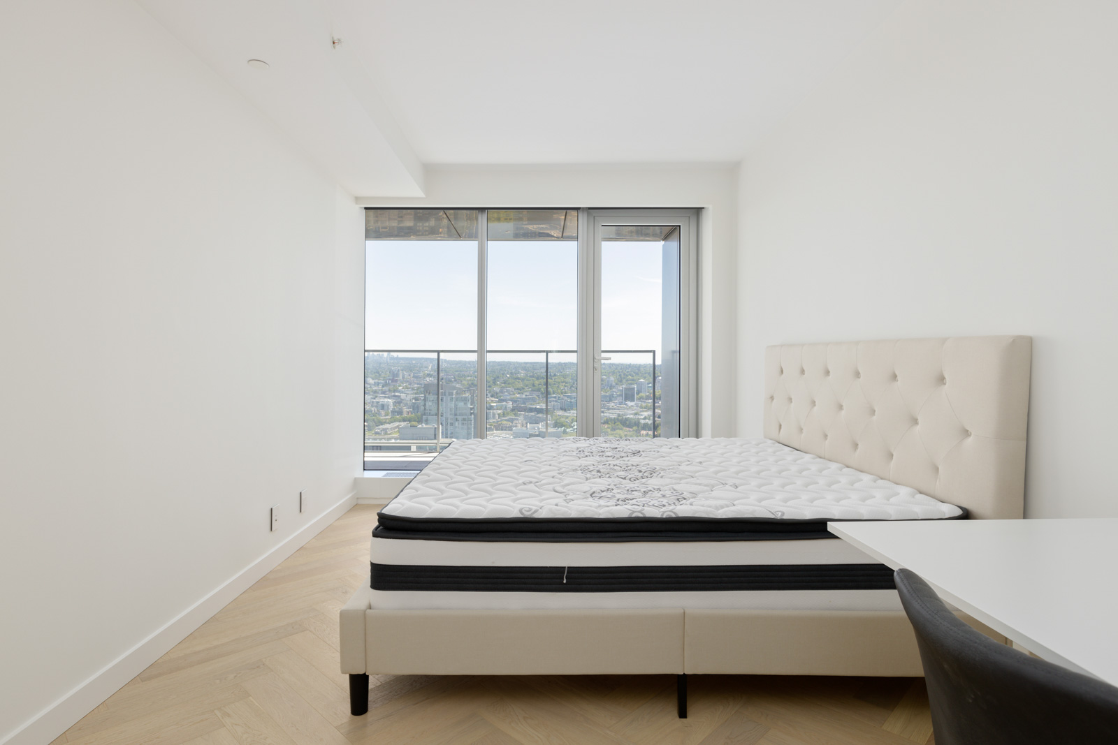 Minimalist bedroom with a beige upholstered bed, mattress, large windows, city view, and light wood flooring. A white desk and black chair are visible in the foreground.
