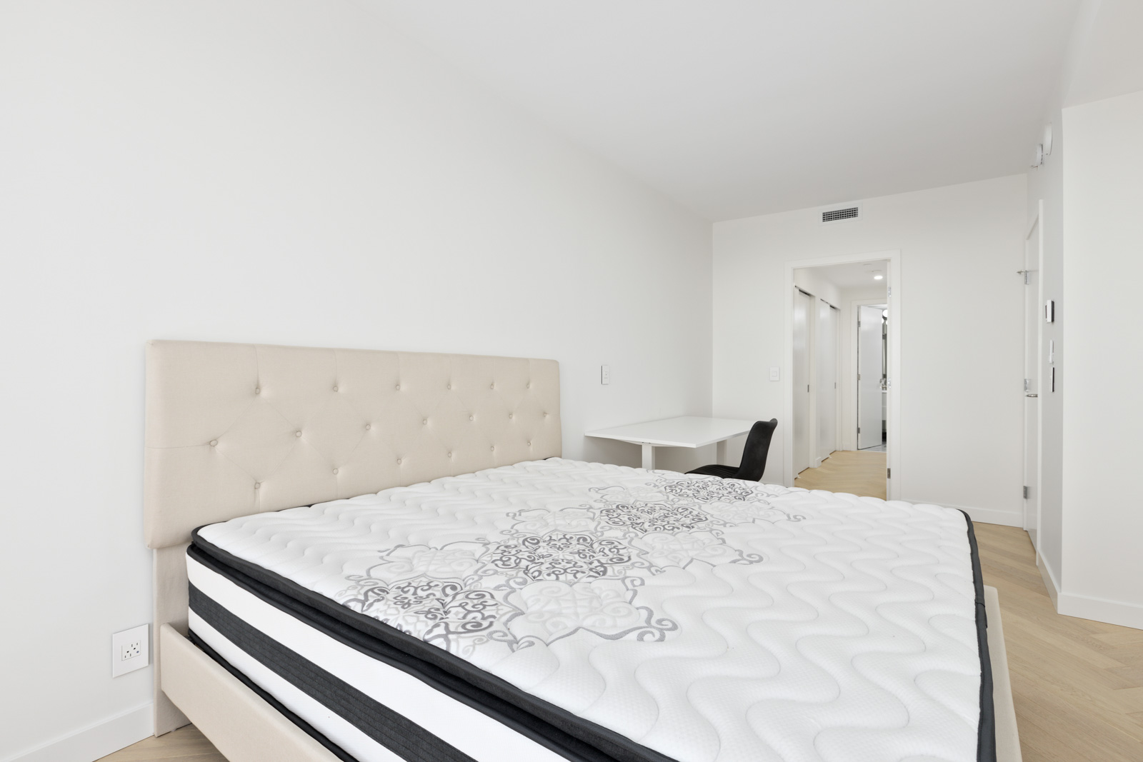 Minimalist bedroom with a beige tufted headboard, white mattress, small white desk, and light wood flooring; walls and decor are white.