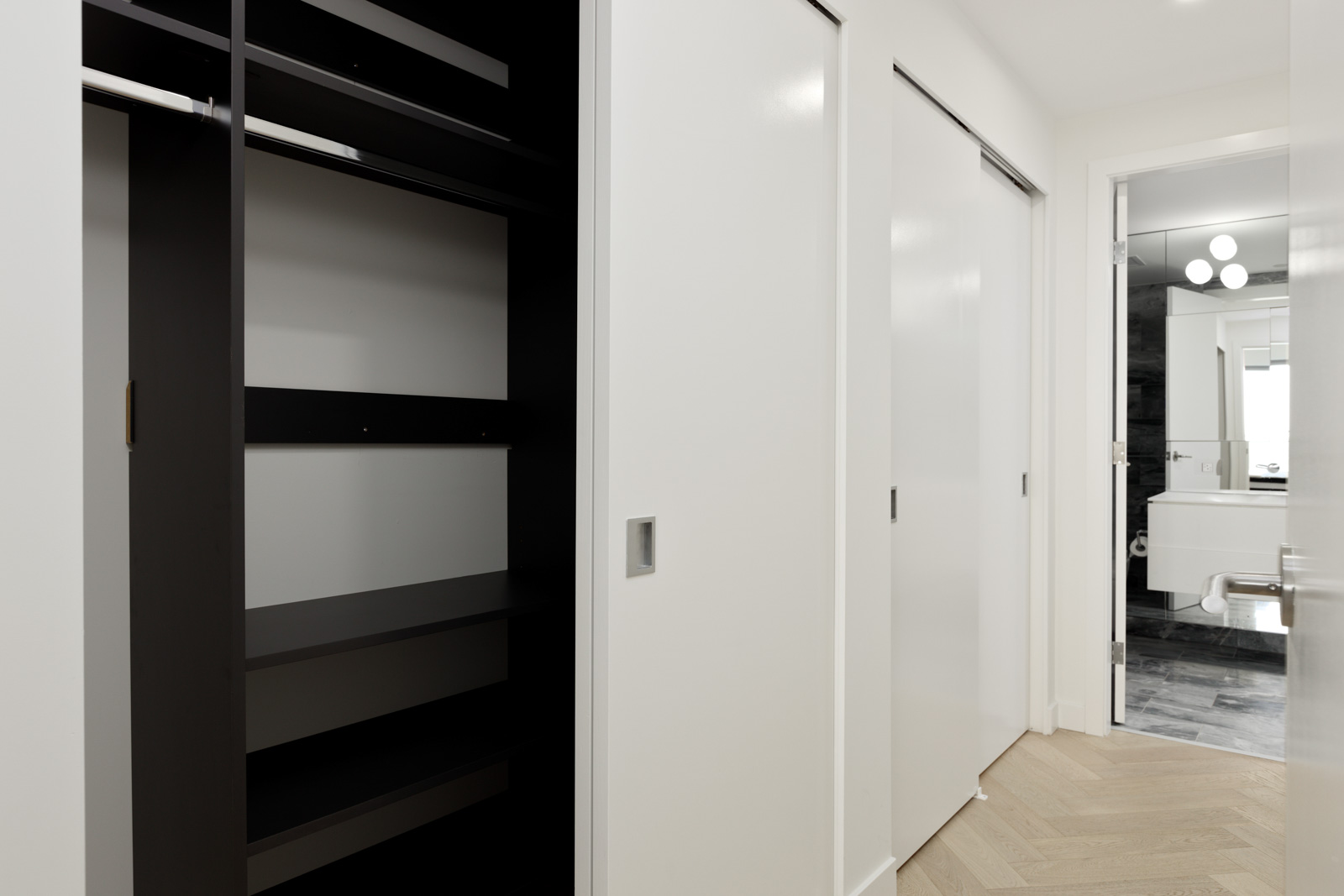 Empty closet with open shelving and hanging rod next to a hallway with white sliding doors, leading to a modern bathroom with a sink, mirror, and round light fixtures.