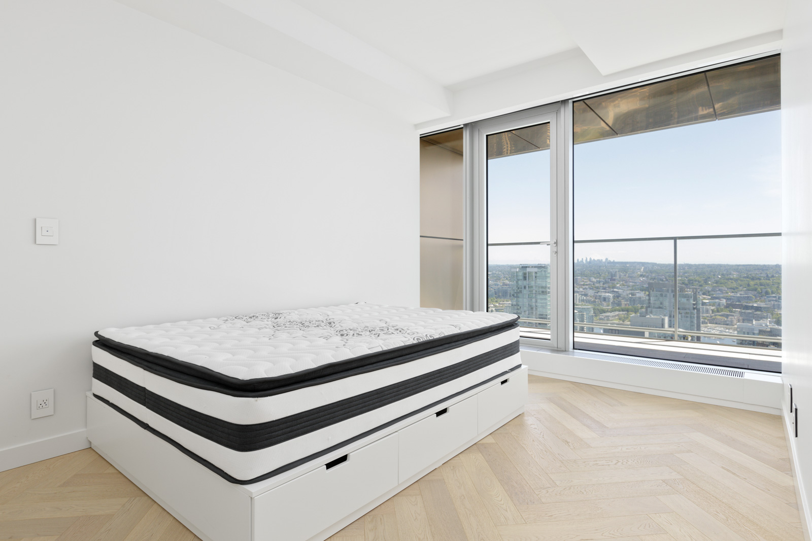 Modern bedroom with a bed featuring built-in storage drawers, large windows, city view, and light wood herringbone flooring.
